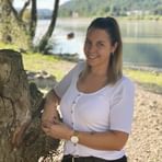 Anja Bodenhofer in weißer Strickjacke lehnt an verwittertem Baumstumpf am ruhigen See mit Hügeln im Hintergrund an sonnigem Tag.