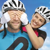 Zwei lächelnde Radfahrer mit weißen Uvex-Helmen und Radkleidung. Frau hält dem Mann spielerisch die Augen zu vor blauem Himmel mit Wolken.