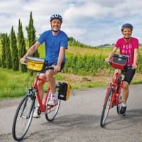 Two smiling cyclists with helmets on touring bikes on a country road in Tuscany, surrounded by vineyards and tall cypress trees.