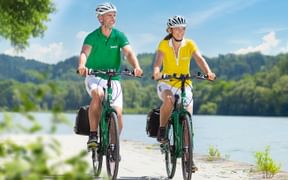 Mann in grünem Shirt und Frau in gelber Weste radeln auf gepflastertem Weg am See. Beide tragen Helme und Packtaschen. Hügel und blauer Himmel sichtbar.