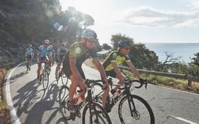 Gruppe von Radfahrern auf einer Küstenstraße mit Meerblick. Zwei Radfahrer in gelben Trikots führen, gefolgt von anderen in blauen Trikots.