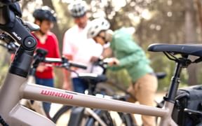 Nahaufnahme eines Winora E-Bike-Rahmens im Vordergrund mit unscharfer Radfahrergruppe mit Helmen in Waldumgebung.