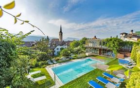 Außenpool im Hotel Goldener Stern in Kaltern mit blauen Liegen, umgeben von grünem Rasen und Bäumen. Kirchturm im Hintergrund sichtbar.
