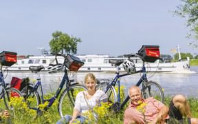 Drei Radfahrer entspannen in einer Blumenwiese am Fluss mit Fahrrädern und einem weißen Kreuzfahrtschiff im Hintergrund unter blauem Himmel.