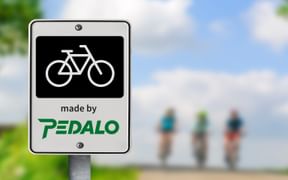 Weißes Schild mit schwarzem Fahrradsymbol und grünem Pedalo-Logo an einem Pfosten. Verschwommene Radfahrer auf Weg im Hintergrund unter blauem Himmel.