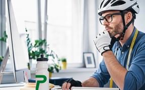 Mann mit Fahrradhelm und Brille am Schreibtisch mit Computer, Wasserflasche und 'Tailor Made'-Schild, plant maßgeschneiderte Radreisen.