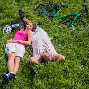 Zwei Radfahrer in Pedalo-Radbekleidung liegen auf grünem Gras. Eine Frau im rosa Trikot und eine Person im hellvioletten Shirt ruhen neben einem grünen Fahrrad.