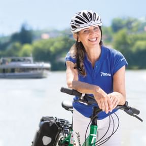 Frau in blauem Pedalo-Radtrikot und weißem Helm auf grünem Fahrrad am Fluss. Ein Kreuzfahrtschiff ist im Hintergrund auf dem Wasser sichtbar.