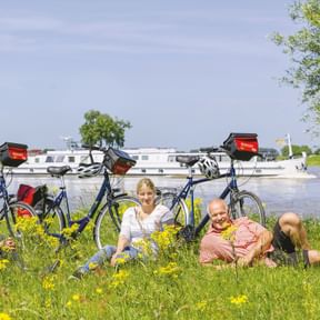 Drei Radfahrer entspannen in einer Blumenwiese am Fluss mit Fahrrädern und einem weißen Kreuzfahrtschiff im Hintergrund unter blauem Himmel.