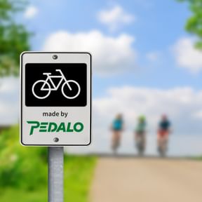 Weißes Schild mit schwarzem Fahrradsymbol und grünem Pedalo-Logo an einem Pfosten. Verschwommene Radfahrer auf Weg im Hintergrund unter blauem Himmel.