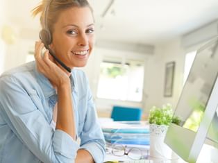 Blonde woman wearing headset smiling at camera while working at desk with computer monitor, glasses, and plant in bright office.