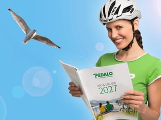 Lächelnde Frau mit weißem Fahrradhelm und grünem Shirt hält Pedalo Katalog 2027 vor blauem Himmel mit fliegender Möwe.