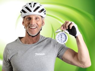 Lächelnder Radfahrer mit weißem Helm und grauem Shirt hält weißen Wecker. Grüner kreisförmiger Hintergrund hinter ihm.