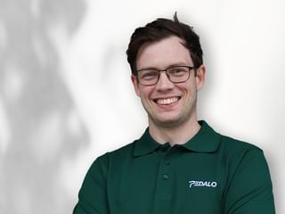 Portrait of Sandi wearing glasses and a green Pedalo polo shirt, smiling at the camera against a white background.