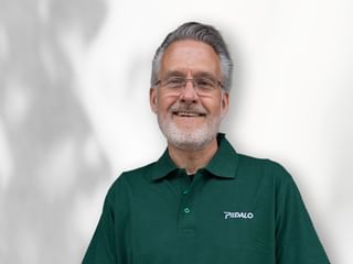 Portrait eines lächelnden Mannes mit Brille und grauen Haaren in einem dunkelgrünen Pedalo-Poloshirt vor weißem Hintergrund.