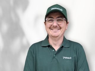 Porträt von Nicolas Diksaitis mit Brille, grüner Pedalo-Kappe und Poloshirt, lächelnd vor weißem Hintergrund.