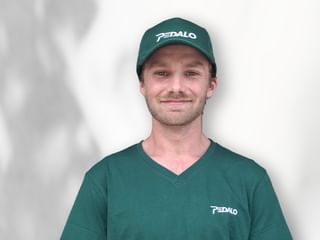 Porträt von Aidan Munn mit grüner Pedalo-Kappe und passendem V-Ausschnitt-Shirt mit Pedalo-Logo, lächelnd vor weißem Hintergrund.