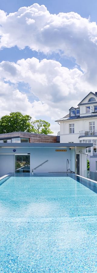 Dach-Infinity-Pool der Villa Hügel Trier mit Glasgeländer, Liegestühlen und weißem Villengebäude unter dramatischem Wolkenhimmel.