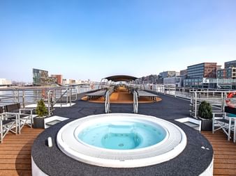 Oberdeck des Schiffs Swiss Crown mit rundem weißen Whirlpool, weißen Sitzbereichen und moderner Stadtsilhouette im Hintergrund.