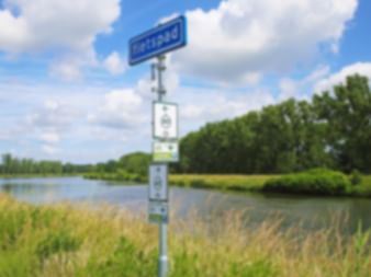 Weißer Wegweiser mit mehreren Richtungsschildern neben einem ruhigen Fluss. Grüne Bäume säumen das gegenüberliegende Ufer unter blauem Himmel.