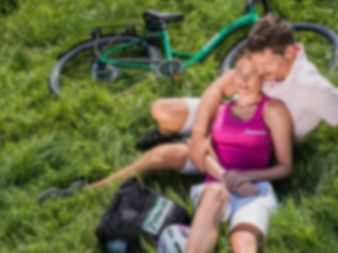 Paar ruht auf grünem Gras mit einem türkisfarbenen Fahrrad im Hintergrund. Die Frau trägt ein pinkes Oberteil, der Mann ein weißes Hemd.