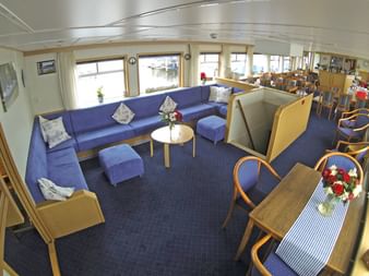 Geräumige Schiffslounge mit blauen Sitzgelegenheiten, Holztischen und großen Fenstern. Mit L-förmigem Sofa, Essbereich und Hafenblick.