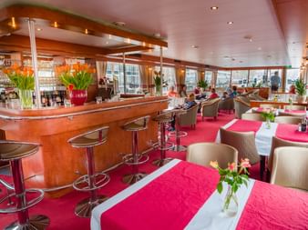 Innenraum des Fluvius Schiffrestaurants mit Holzbar mit orangen Tulpen, Esstischen mit roten Tischdecken, beigen Stühlen und Panoramafenstern.