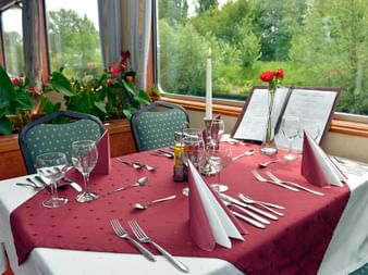 Eleganter Esstisch an Bord des Fluvius Schiffs mit roter Tischdecke, Weingläsern, Kerze und roten Blumen. Grüne Polsterstühle vor Fenstern.