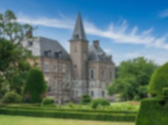 Mittelalterliches Schloss mit spitzem Turm und mehreren Gebäuden, umgeben von gepflegten Gärten und grünen Rasenflächen unter blauem Himmel.