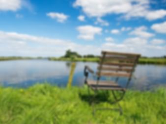 Holzstuhl auf grünem Gras neben einem ruhigen See mit blauem Himmel und weißen Wolken, die sich im Wasser spiegeln.