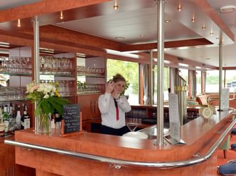 Innenraum der Schiff Fluvius Bar mit weiblicher Barkeeperin in weißem Hemd hinter gebogener Holztheke. Glasregale, Flaschen und Blumenvase sichtbar.