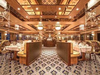 Luxuriöser mehrstöckiger Speisesaal an Bord der Swiss Crown mit verzierter Decke, gemustertem Teppich, weiß gedeckten Tischen und eleganten Leuchten.