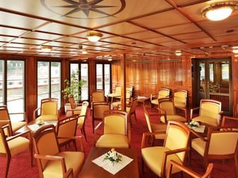 Elegante Schiffslounge mit gelben Lederstühlen, rotem Teppich, Holzvertäfelung und großen Fenstern mit Wasserblick auf der Florentina.