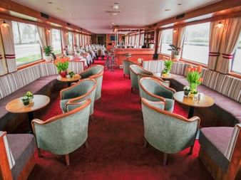 Luxuriöse Schiffslounge mit grünen Ledersesseln, rotem Teppich, gestreiften Sitzbänken und großen Fenstern. Barbereich im Hintergrund.