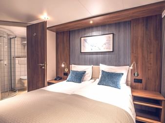 Moderne Schiffskabine auf der Swiss Crown mit Doppelbett, holzgetäfelten Wänden, blauen Kissen und sichtbarem Badezimmer mit Glasduschtür.