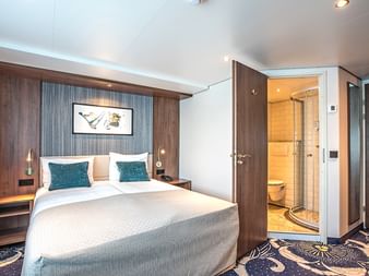 Moderne Schiffskabine auf der Swiss Crown mit Doppelbett, dunkler Holzvertäfelung, türkisfarbenen Kissen und verglastem Bad mit Dusche.
