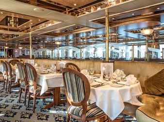 Luxuriöser Speisesaal auf dem Schiff Swiss Crown mit weißen Tischdecken, gestreiften Polsterstühlen, goldenen Banksitzen und großen Fenstern.