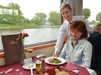 Kellnerin serviert Essen an ältere Passagierin an Bord des Fluvius Schiffs. Tisch gedeckt mit Menü, Blumen und Getränken mit Flussblick.