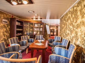 Elegante Schiffsbibliothek-Lounge auf der Florentina mit blauen gemusterten Sesseln, Holztisch, Bücherregalen, goldener Tapete und rotem Teppich.