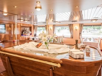 Luxuriöser Bootsinnenraum mit geschwungener Holzbar, weißen Blumen, Essbereich mit beigen Sitzen und großen Fenstern mit Wasserblick.