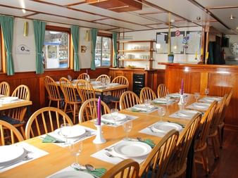 Innenraum des Flora Schiff Speisesaals mit Holzstühlen um Tische mit weißen Tellern, Gläsern und Servietten. Grüne Vorhänge rahmen Fenster.