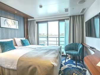 Moderne Schiffskabine auf der Swiss Crown mit Doppelbett, türkisfarbenem Stuhl, Wandfernseher und Glastür zum Balkon mit Wasserblick.