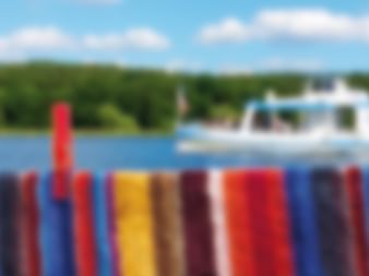 Bunte gestreifte Reling im Vordergrund mit weißem Flusskreuzfahrtschiff auf blauem Wasser. Grüne Hügel und blauer Himmel im Hintergrund.