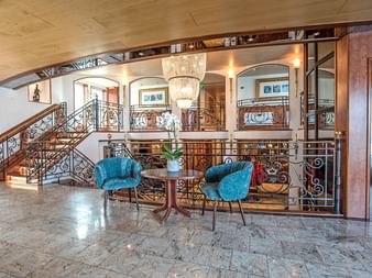Luxuriöses Schiffsinterieur mit Marmorböden, verzierten Eisengeländern, Kristallleuchter und zwei türkisfarbenen Sesseln um einen kleinen Tisch.