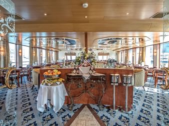 Eleganter Speisesaal an Bord der Swiss Crown mit verzierten Messinggeländern, weiß gedeckten Tischen, dekorativem Teppich und großen Fenstern.