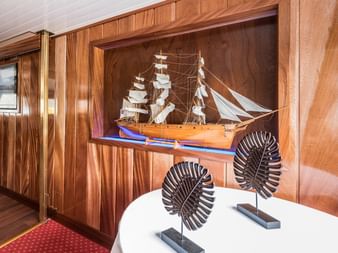 Innenraum des Schiffs Florentina mit holzgetäfelten Wänden und eingebauter Vitrine mit detailliertem Segelschiffmodell. Weißer Tisch mit Dekoobjekten im Vordergrund.