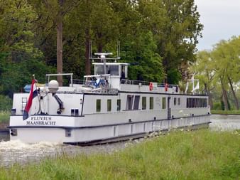 Weißes Flusskreuzfahrtschiff Fluvius aus Maasbracht fährt durch einen schmalen Kanal, umgeben von üppigen grünen Bäumen und Vegetation.