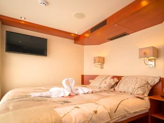 Elegante Schiffskabine mit Doppelbett, weißer Bettwäsche, dekorativem Handtuchschwan, Wandfernseher und warmen Holzakzenten mit Ambientebeleuchtung.