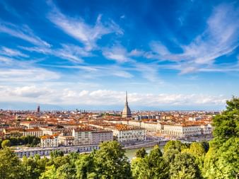 Panoramablick auf Turin mit dem ikonischen Mole Antonelliana Turm über rotgedeckten Gebäuden unter dramatischem blauen Himmel mit weißen Wolken.