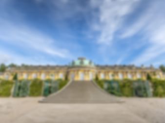 Gelbes Schloss mit symmetrischer Fassade und grünem Kupferdach. Terrassierte Weingärten mit geschnittenen Hecken führen zum Gebäude unter blauem Himmel.